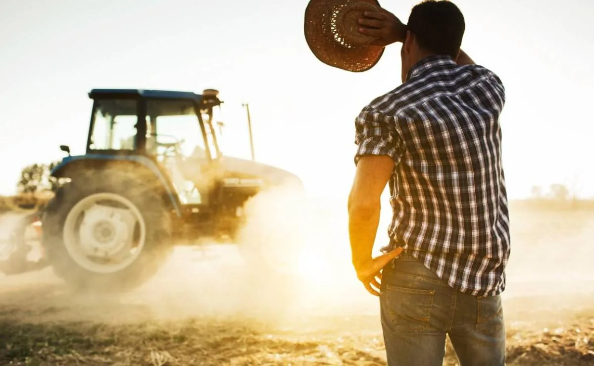 Agricultor de espaldas observando un tractor en funcionamiento levantando polvo en un campo agrícola durante el atardecer.