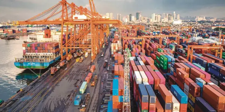 Vista aérea de un puerto comercial con un barco de carga, grúas naranjas y contenedores apilados.