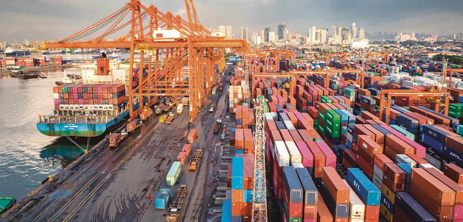 Vista aérea de un puerto comercial con un barco de carga, grúas naranjas y contenedores apilados.