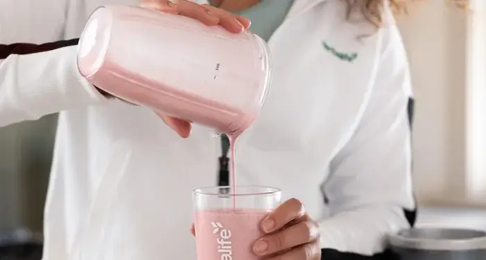 Mujer sonriente vierte batido rosa de Herbalife en un vaso en la cocina. Promueve bienestar y energía para un estilo de vida activo.