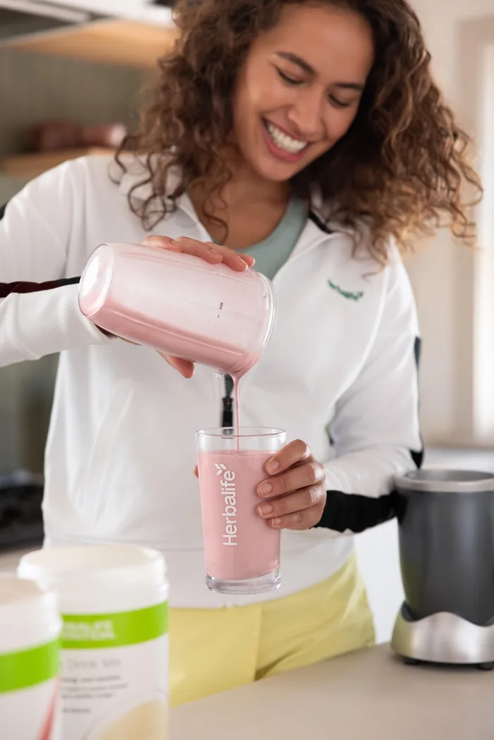 Mujer sonriente vierte batido rosa de Herbalife en un vaso en la cocina. Promueve bienestar y energía para un estilo de vida activo.
