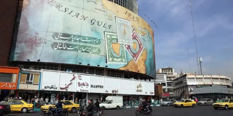Gran cartel publicitario con mapa del Golfo Pérsico, sellos postales y texto en persa, sobre edificios comerciales y calle con tráfico vehicular.