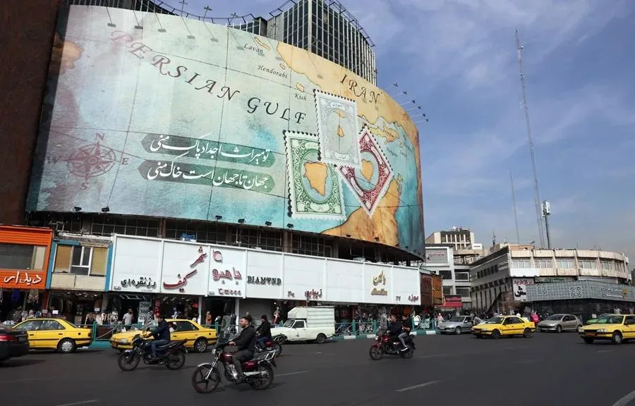 Gran cartel publicitario con mapa del Golfo Pérsico, sellos postales y texto en persa, sobre edificios comerciales y calle con tráfico vehicular.