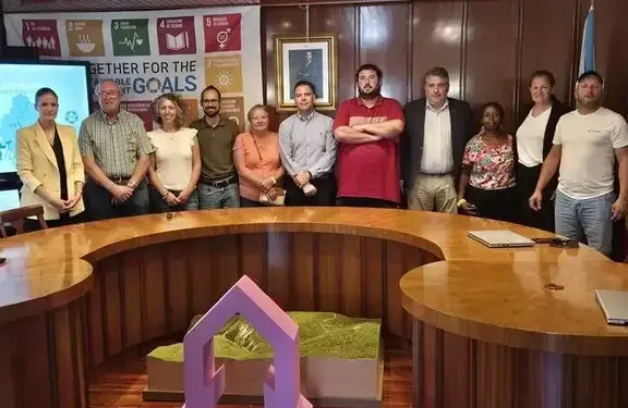 Grupo diverso de personas posando juntas frente a un cartel de los Objetivos de Desarrollo Sostenible de la ONU y una mesa de conferencias.