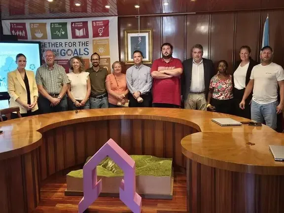Grupo diverso de personas posando juntas frente a un cartel de los Objetivos de Desarrollo Sostenible de la ONU y una mesa de conferencias.