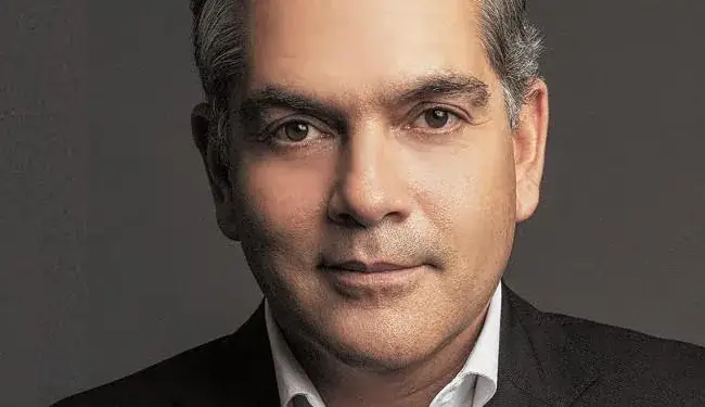 Primer plano de un hombre profesional con traje oscuro y camisa blanca, cabello grisáceo, mirando al frente con expresión confiada.
