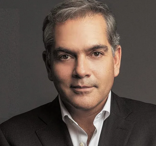 Primer plano de un hombre profesional con traje oscuro y camisa blanca, cabello grisáceo, mirando al frente con expresión confiada.