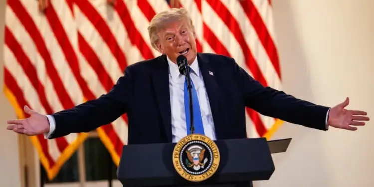 Donald Trump con traje oscuro y corbata azul hablando detrás de un atril con el sello presidencial, flanqueado por banderas de Estados Unidos.