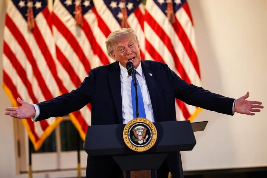 Donald Trump con traje oscuro y corbata azul hablando detrás de un atril con el sello presidencial, flanqueado por banderas de Estados Unidos.