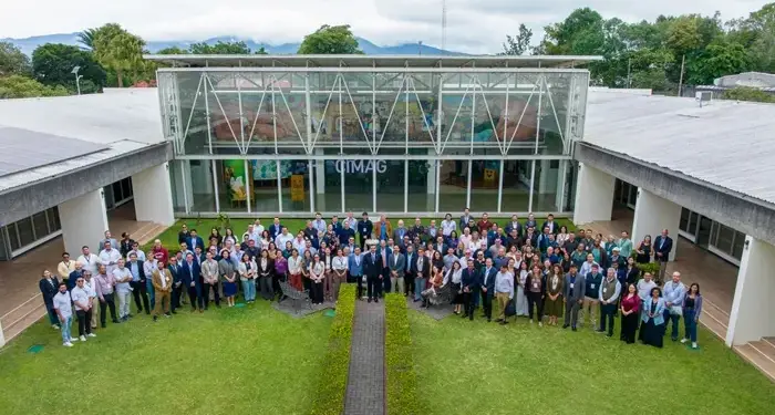 Gran grupo de profesionales diversos posando en un césped frente a un edificio moderno con fachada de cristal y paneles solares. Ambiente de evento corporativo o conferencia.