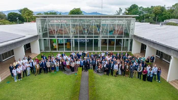 Gran grupo de profesionales diversos posando en un césped frente a un edificio moderno con fachada de cristal y paneles solares. Ambiente de evento corporativo o conferencia.