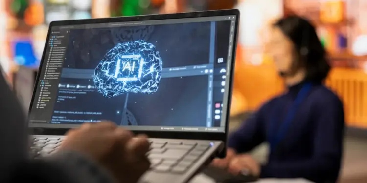 Primer plano de manos escribiendo en un portátil con una imagen de cerebro y 'AI' en pantalla, simbolizando inteligencia artificial y programación. Fondo borroso de oficina.