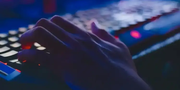 Primer plano de una mano escribiendo sobre un teclado con iluminación azul y roja en un entorno oscuro.