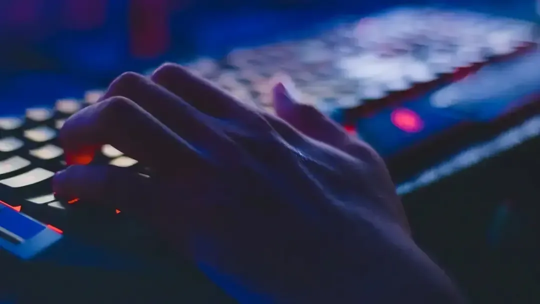 Primer plano de una mano escribiendo sobre un teclado con iluminación azul y roja en un entorno oscuro.
