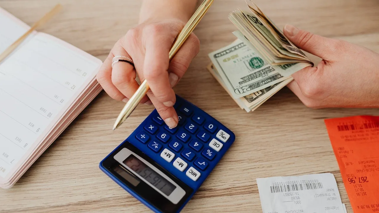 Persona calculando cuentas con billetes en dólares, calculadora azul, notas y recibos sobre una mesa de madera.