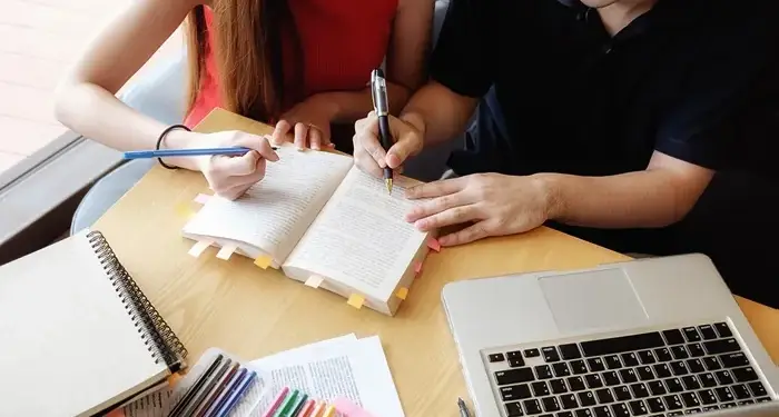 Dos personas revisando un libro con marcadores y una laptop sobre una mesa de madera.