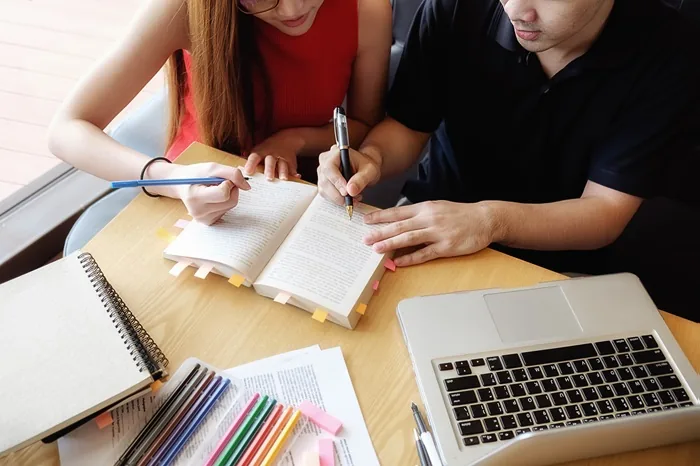 Dos personas revisando un libro con marcadores y una laptop sobre una mesa de madera.