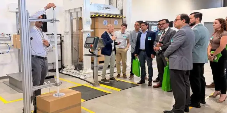 Grupo de profesionales observa una demostración de pruebas de embalaje con un equipo de carga y caída. Un experto explica el proceso junto a cajas de cartón en un entorno industrial.