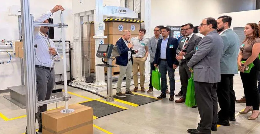 Grupo de profesionales observa una demostración de pruebas de embalaje con un equipo de carga y caída. Un experto explica el proceso junto a cajas de cartón en un entorno industrial.