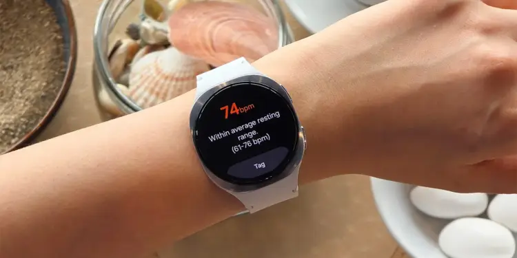 Persona usando un reloj inteligente blanco que muestra una lectura de frecuencia cardíaca de 74 bpm en su pantalla.
