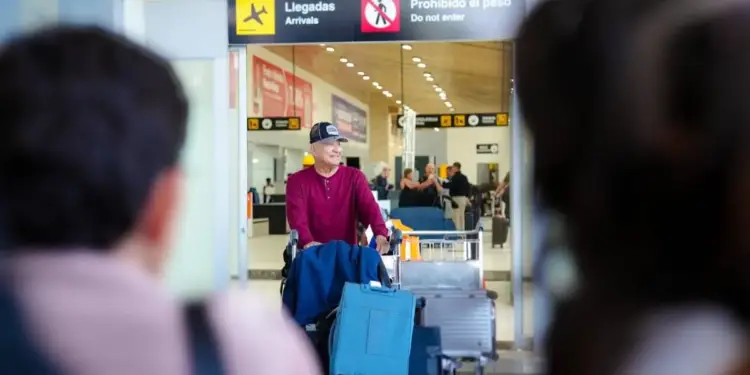Hombre sonriente empujando un carrito con maletas en el área de llegadas de un aeropuerto.