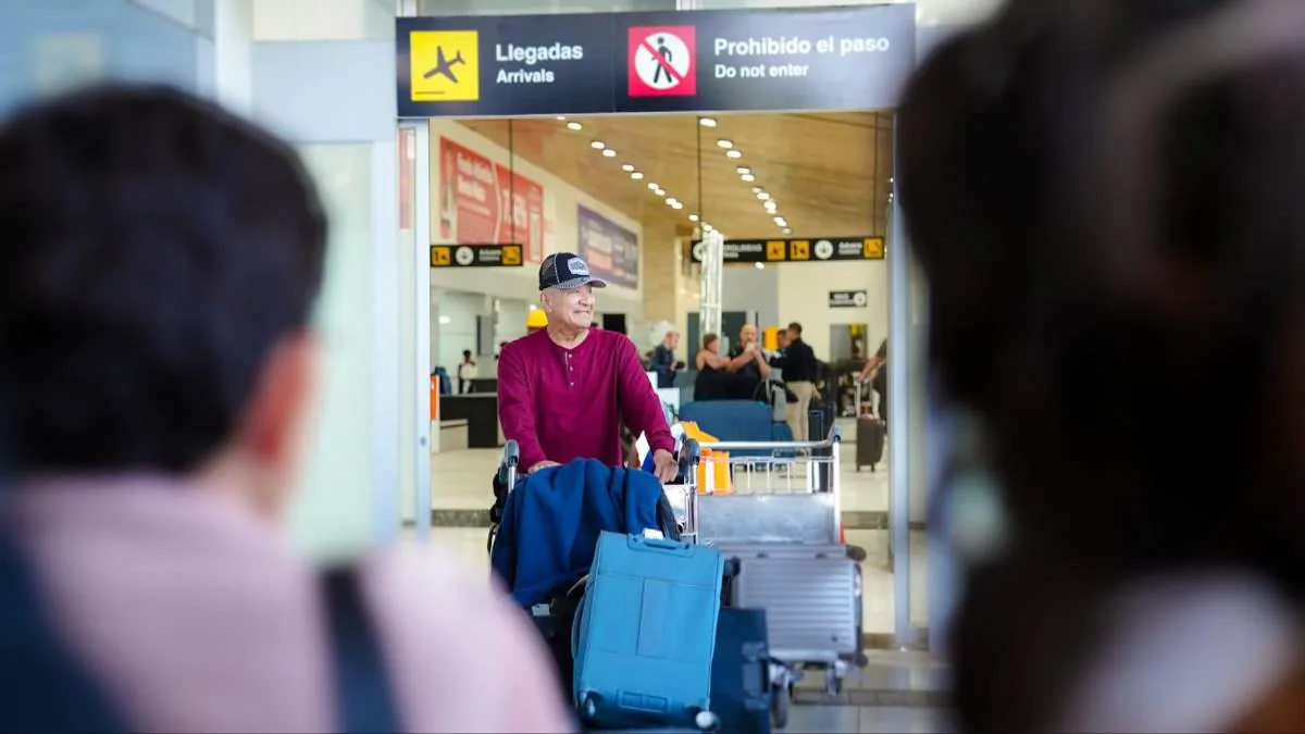 Hombre sonriente empujando un carrito con maletas en el área de llegadas de un aeropuerto.
