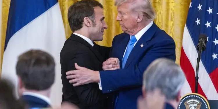 Dos líderes masculinos en trajes formales se saludan frente a las banderas de Francia y Estados Unidos, simbolizando relaciones diplomáticas y económicas.
