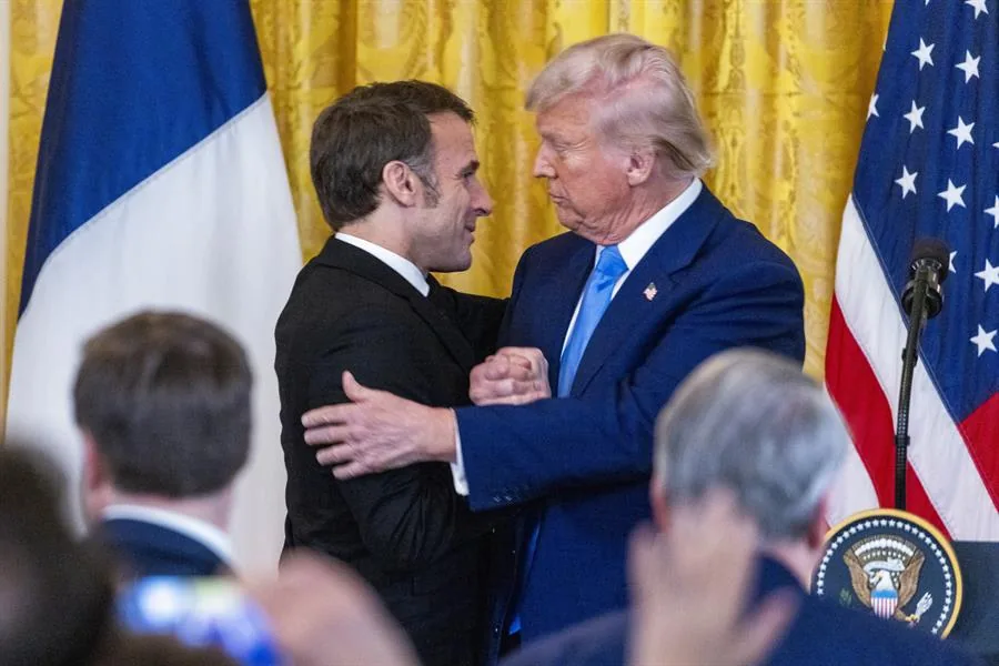 Dos líderes masculinos en trajes formales se saludan frente a las banderas de Francia y Estados Unidos, simbolizando relaciones diplomáticas y económicas.