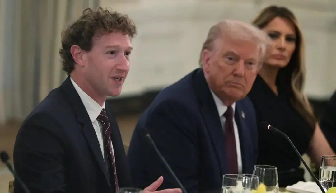 Un hombre con traje y corbata hablando en una reunión formal junto a otros asistentes sentados.