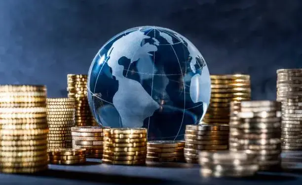 Esfera de cristal con forma de globo terráqueo rodeada por múltiples pilas de monedas doradas sobre fondo oscuro.