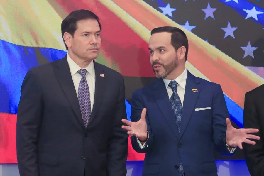 Dos hombres con trajes oscuros y corbatas discuten activamente frente a un fondo con los colores de la bandera de Estados Unidos y Venezuela.