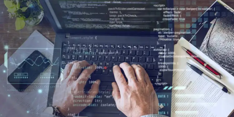 Manos escribiendo en un portátil con código en pantalla. Un smartphone con gráfico de datos, documentos y bolígrafos. Representa análisis tecnológico y financiero.