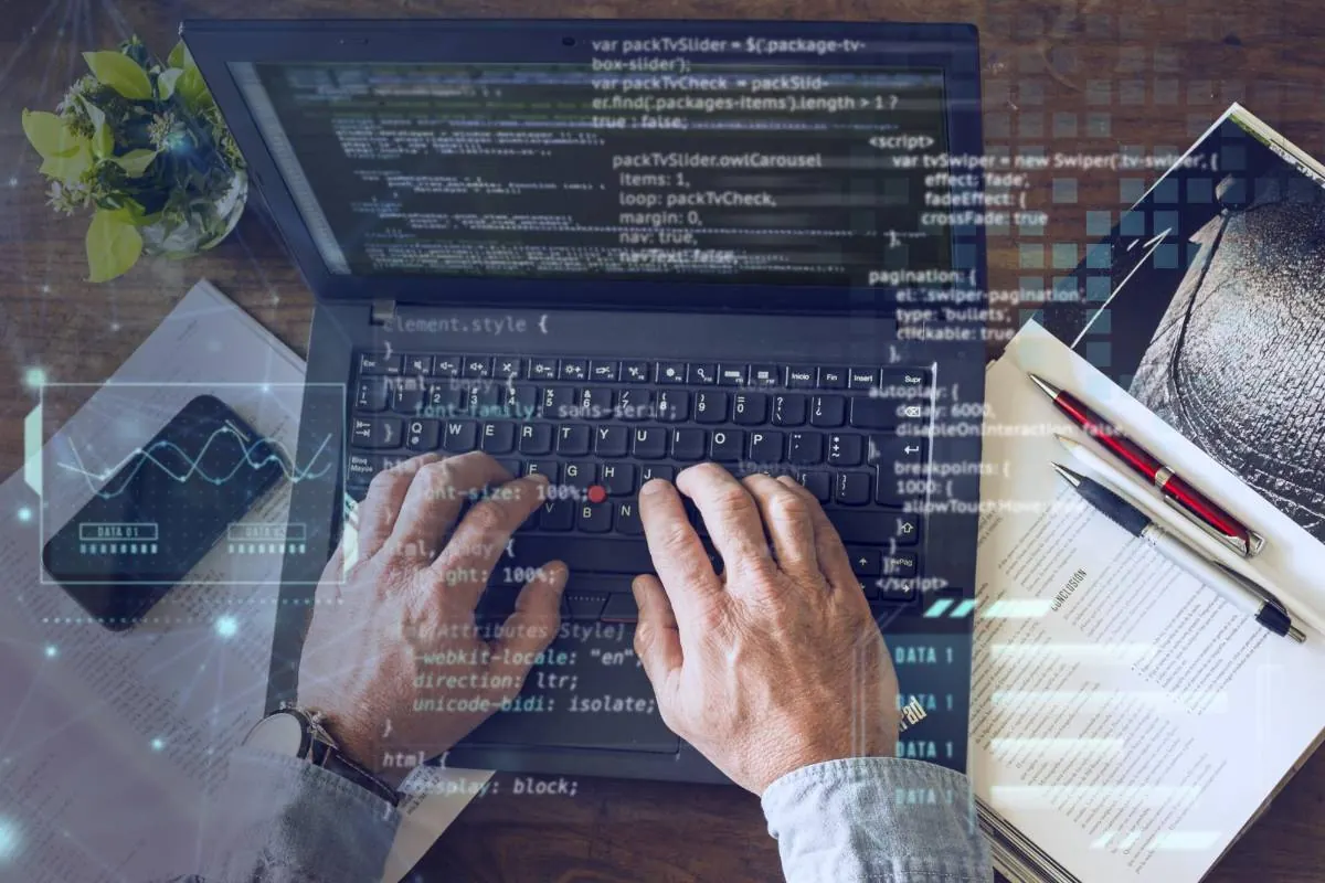 Manos escribiendo en un portátil con código en pantalla. Un smartphone con gráfico de datos, documentos y bolígrafos. Representa análisis tecnológico y financiero.