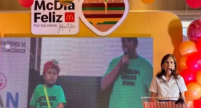 Mujer en podio con logo McDonald's en evento McDía Feliz. Pantalla muestra niño y hombre en escenario con globos y decoración temática.