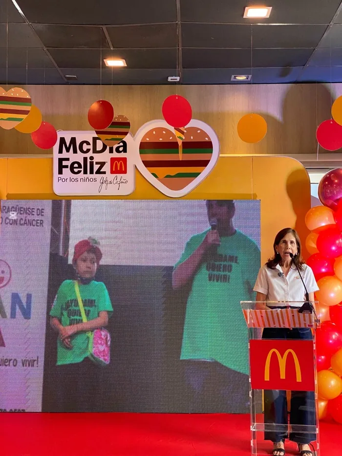 Mujer en podio con logo McDonald's en evento McDía Feliz. Pantalla muestra niño y hombre en escenario con globos y decoración temática.