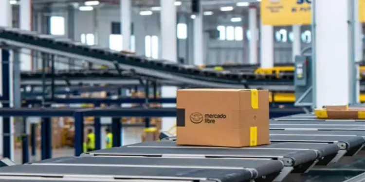 Caja de cartón con el logo de Mercado Libre sobre una cinta transportadora en un centro de distribución logístico.