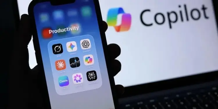Primer plano de un smartphone mostrando una carpeta de aplicaciones de productividad, incluyendo IA como ChatGPT y Gemini. Al fondo, se ve el logo de Copilot en una pantalla.