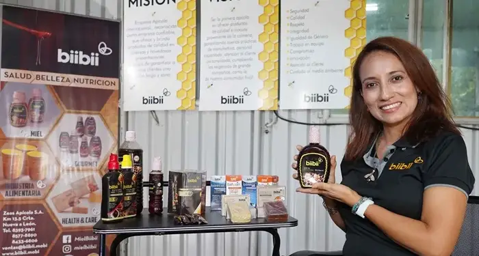 Mujer sonriente sosteniendo una botella de miel frente a una mesa con productos y carteles corporativos de biibii