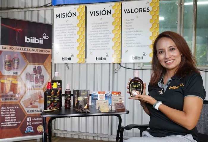 Mujer sonriente sosteniendo una botella de miel frente a una mesa con productos y carteles corporativos de biibii