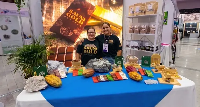 Dos mujeres sonrientes en un stand de feria, exhibiendo productos de cacao y chocolate 'Sauni Gold Bonanza' con vainas y barras.