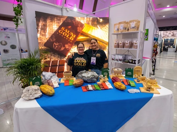 Dos mujeres sonrientes en un stand de feria, exhibiendo productos de cacao y chocolate 'Sauni Gold Bonanza' con vainas y barras.