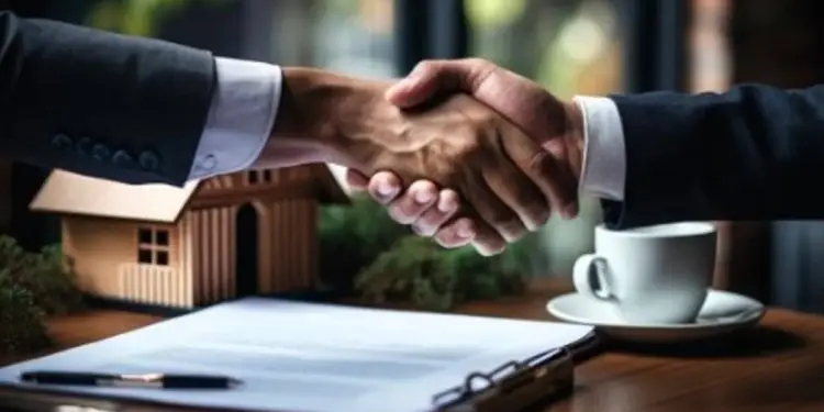 Dos personas con traje se dan la mano sobre una mesa donde hay una maqueta de casa, un contrato y una taza de café.