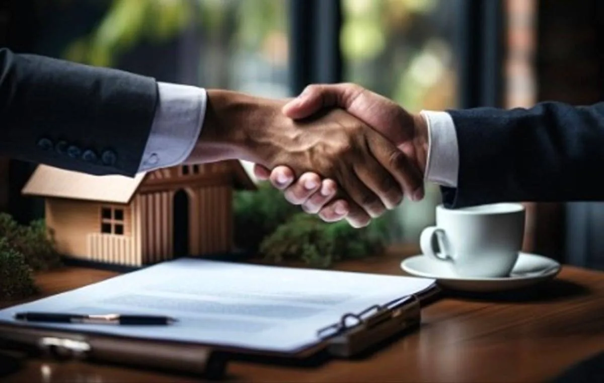 Dos personas con traje se dan la mano sobre una mesa donde hay una maqueta de casa, un contrato y una taza de café.