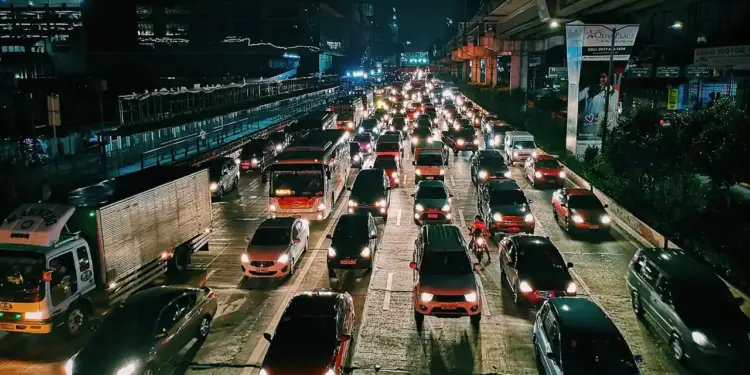 Vista aérea de una autopista urbana con tráfico denso de noche. Numerosos vehículos, incluyendo coches, autobuses y camiones, avanzan lentamente bajo luces brillantes.