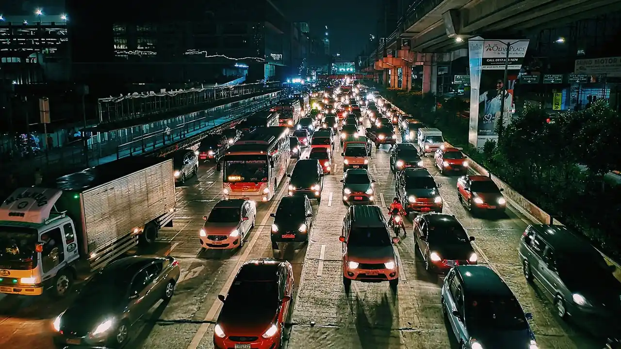 Vista aérea de una autopista urbana con tráfico denso de noche. Numerosos vehículos, incluyendo coches, autobuses y camiones, avanzan lentamente bajo luces brillantes.