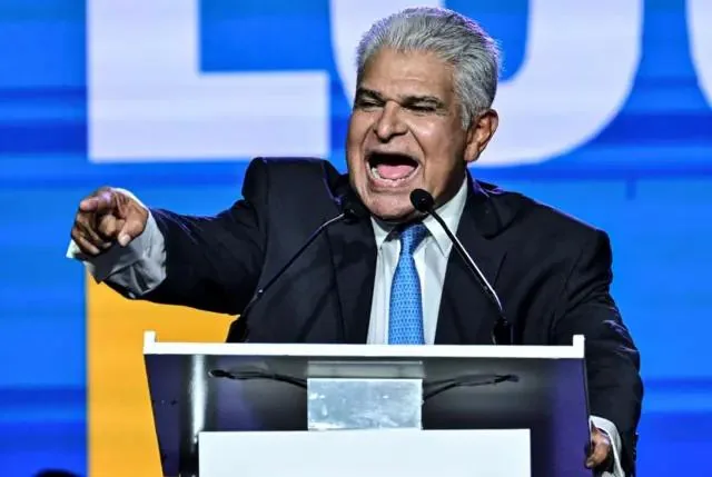 Hombre maduro con traje y corbata hablando apasionadamente frente a un atril en un evento corporativo.