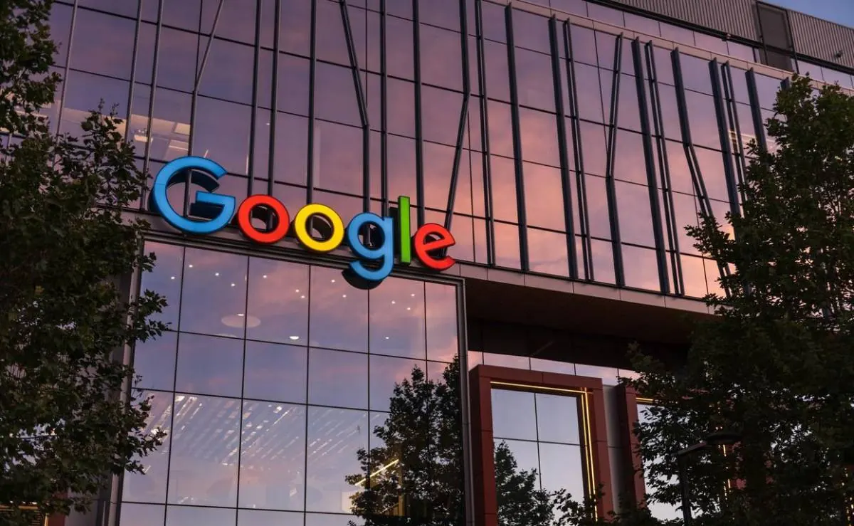 Fachada de cristal de un edificio de oficinas moderno con el logo de Google en colores vibrantes, reflejando un cielo al atardecer.
