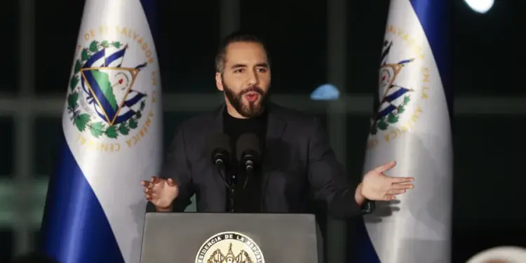 Hombre barbudo en traje oscuro habla en podio con micrófonos, flanqueado por banderas de El Salvador. El podio muestra el escudo presidencial.