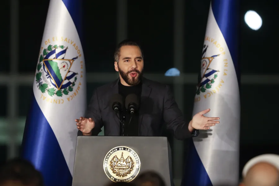 Hombre barbudo en traje oscuro habla en podio con micrófonos, flanqueado por banderas de El Salvador. El podio muestra el escudo presidencial.