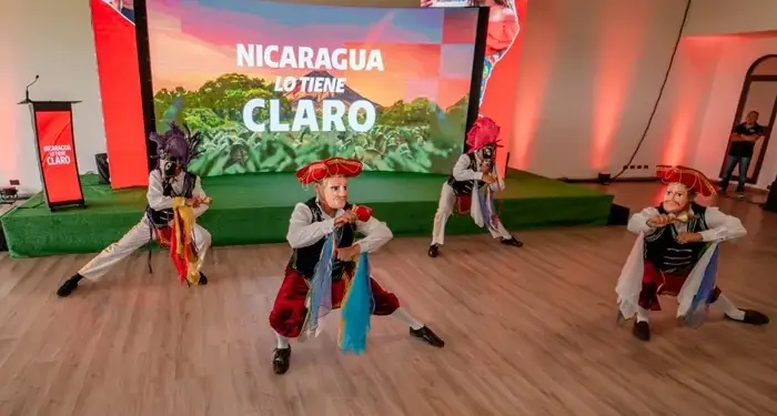 Cuatro bailarines con trajes y máscaras tradicionales actúan en un escenario frente a una pantalla grande con texto.